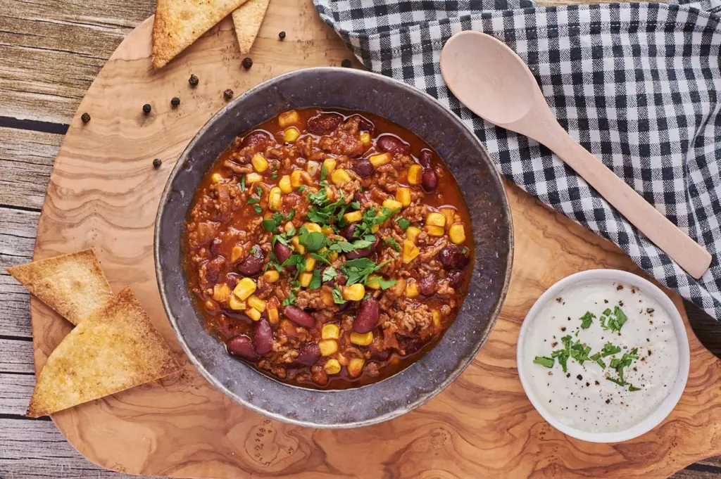 Hausgemachtes Chili con Carne mit Hackfleisch, Mais und Kidneybohnen in einer Schüssel, daneben Tortilla-Ecken und Dip