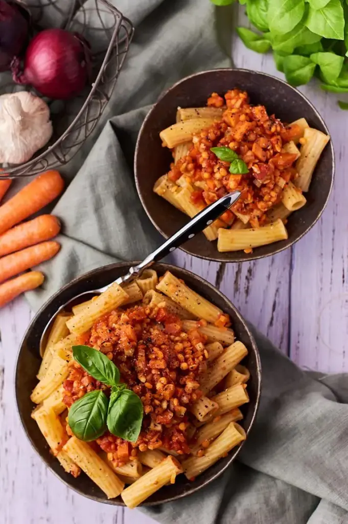 Pasta mit Linsenbolognese und frischem Basilikum, daneben Möhren und Zwiebeln