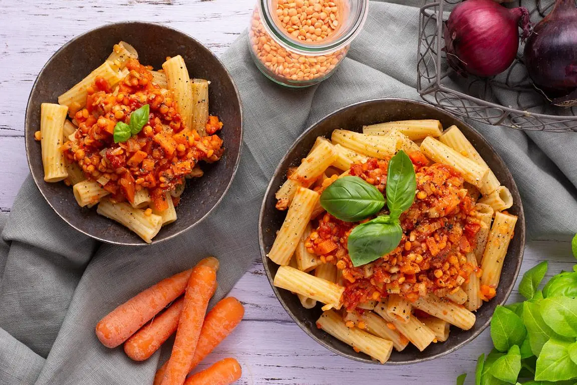 Zwei Schalen Pasta mit Linsenbolognese, daneben Möhren, rote Zwiebeln und Linsen