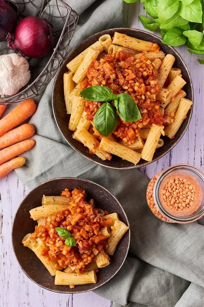 Zwei Portionen vegane Linsenbolognese mit Pasta, umgeben von frischem Gemüse