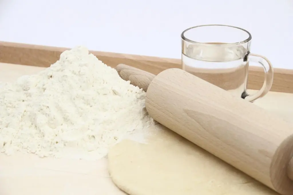 Ein Haufen Mehl liegt auf einer Holzplatte neben einem Nudelholz, einem Glas Wasser und einem bereits ausgerollten Teigstück.