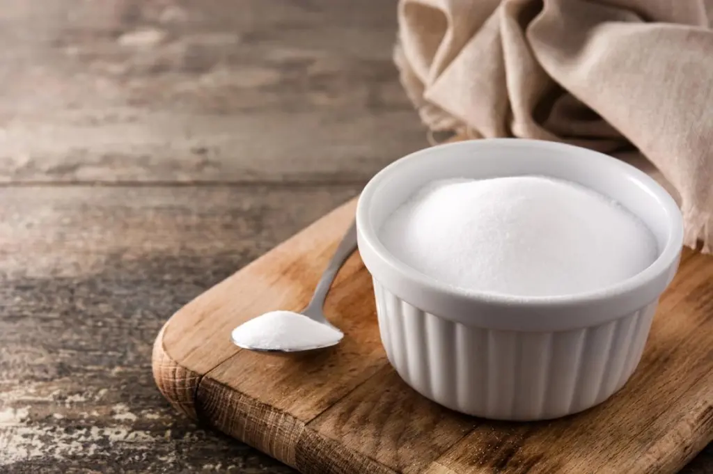 Eine kleine weiße Keramikschale, gefüllt mit feinem Salz, steht mit einem Löffel auf einem rustikalen Holzbrett.