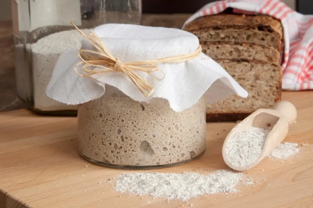 Sauerteig-Starter im Glas mit Stoffabdeckung und Haushaltsgummi neben aufgeschnittenem Sauerteigbrot und Mehl auf Holzbrett