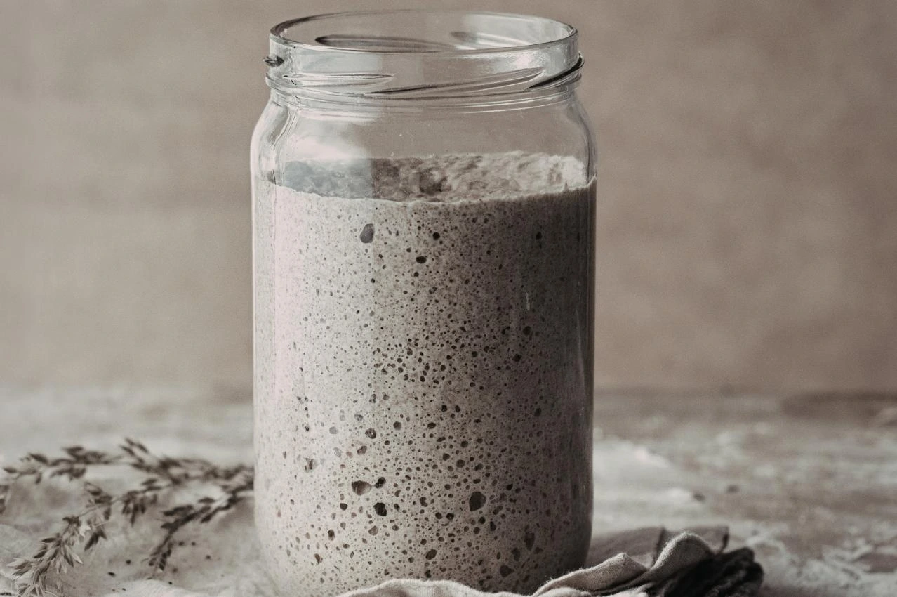 Verdoppelter Sauerteig-Starter voller Blasen im Schraubglas auf Holzbrett mit Getreideähren, backbereit und aktiv