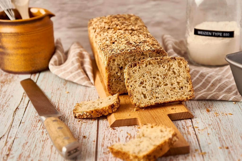 Angeschnittenes Dinkelbrot auf einem Holzbrett mit Brotmesser, am Bildrand die Kastenform