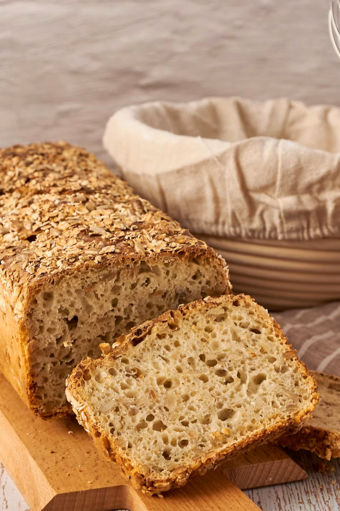 Scheibe Dinkelbrot über Nacht mit saftiger Krume und Sonnenblumenkernen vor dem angeschnittenen Laib