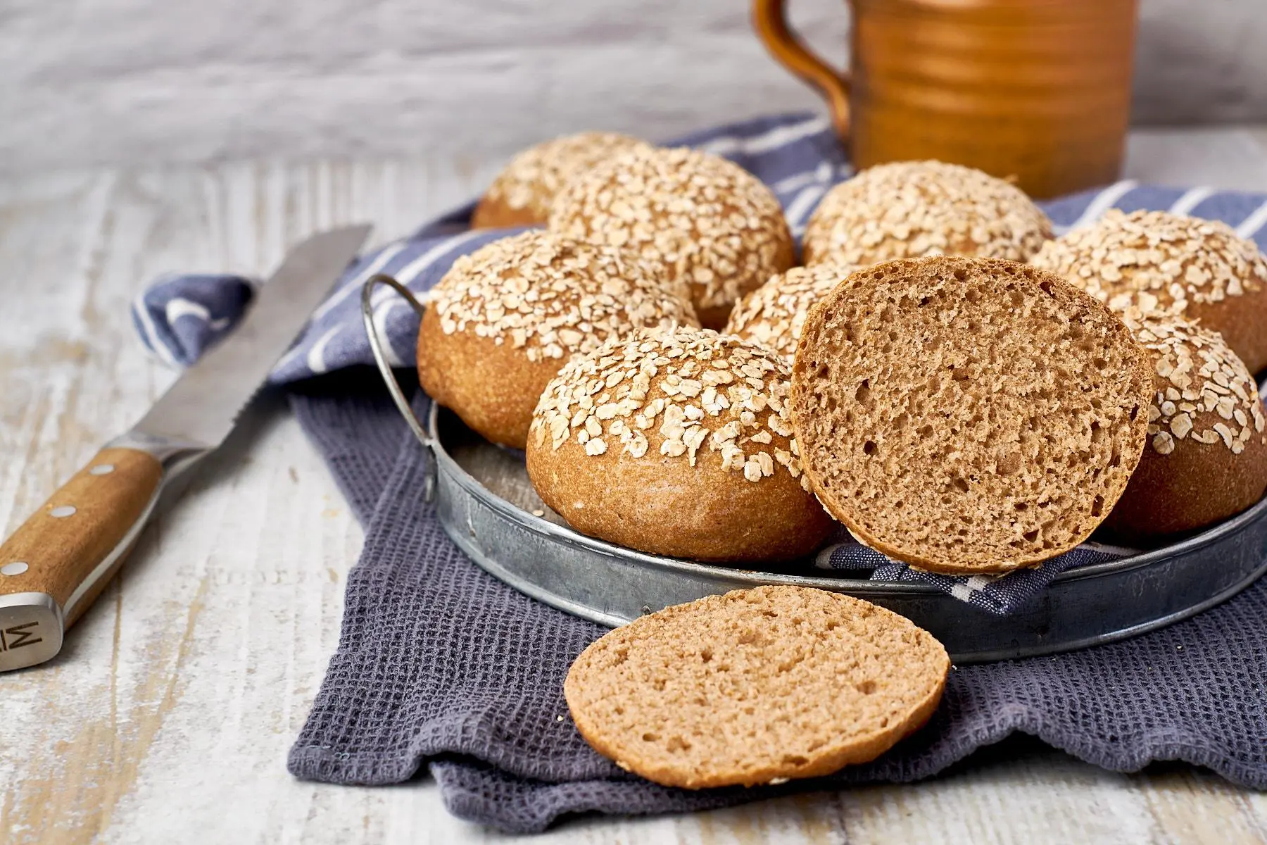 Frühstücksszenerie mit Dinkel-Vollkornbrötchen mit Haferflocken, aufgeschnitten auf Metallteller mit Keramiktasse im Hintergrund