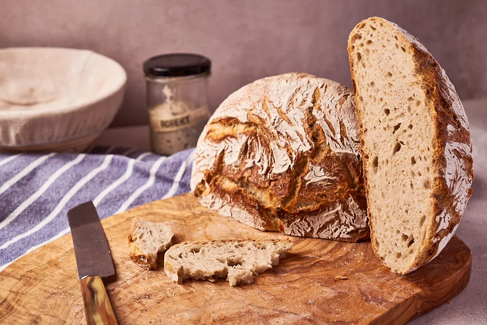 Dinkel-Roggen-Brot mit Sauerteig neben Gärkorb und Roggenstarter im Glas